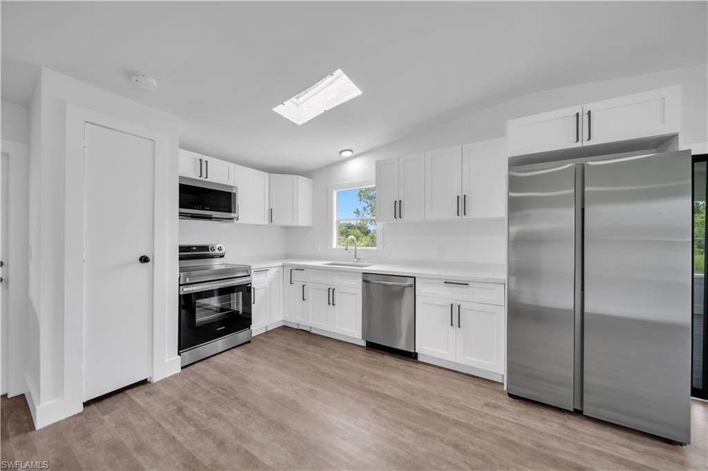 Modern kitchen featuring white cabinetry, stainless steel appliances, a white solid surface countertop, wood-finish flooring, and a ceiling-mounted skylight