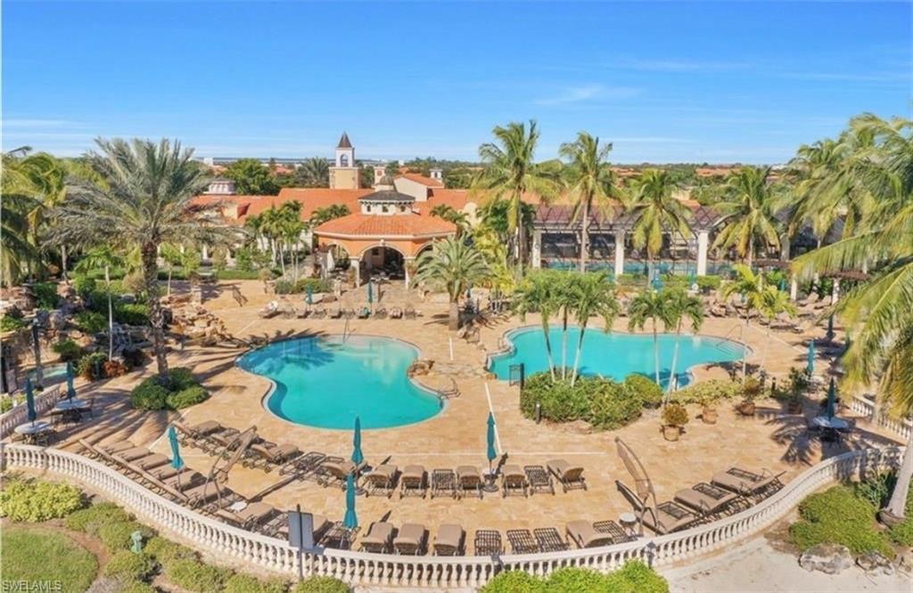 Resort-style resort amenity area featuring two sparkling lagoon-form pools with integrated spa, abundant lounge seating with umbrellas, mature palm tree landscaping, and a Mediterranean-inspired clubhouse with terracotta tile roofing and arched architectural details, all enclosed within decorative perimeter fencing