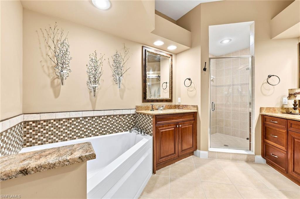 Spa-inspired primary bathroom featuring a soaking tub with decorative tile surround, dual wood-finish vanities with granite countertops, a frameless glass-enclosed shower with tile detailing, recessed lighting, and decorative wall art