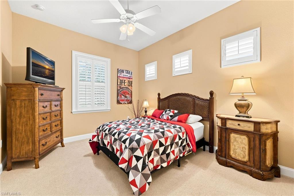 Spacious bedroom featuring warm neutral walls, ceiling fan with integrated lighting, and wood-finish furnishings including a nightstand with brass-accented lamp