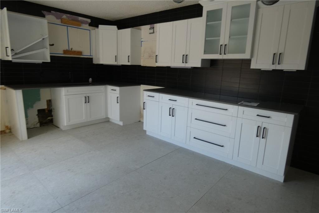 Kitchen featuring white shaker cabinetry, dark countertops, a dark subway tile backsplash, light-toned tile flooring, and integrated display cabinets with frosted glass panels