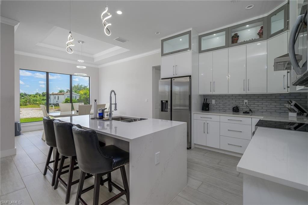 Gourmet kitchen featuring a large waterfall island with integrated sink, stainless steel appliances, white cabinetry with display uppers, and light-toned tile flooring
