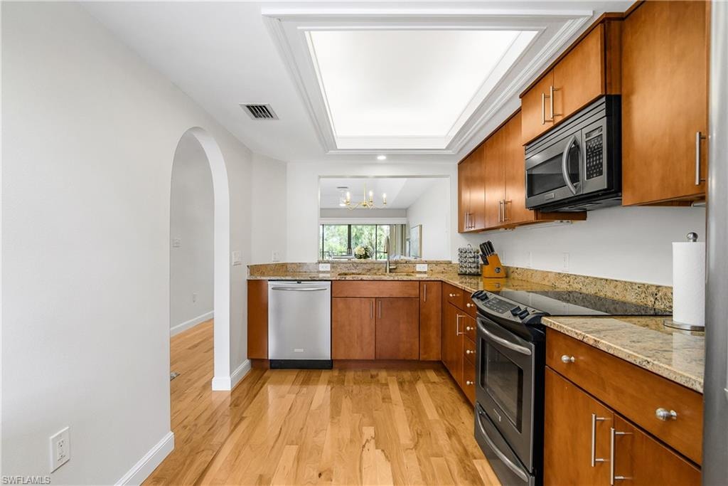Kitchen with brown cabinets, stainless steel appliances, light wood finished floors, light stone countertops, and a chandelier