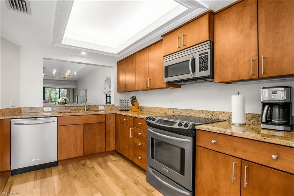 Kitchen with appliances with stainless steel finishes, brown cabinetry, a chandelier, light wood-type flooring, and light stone counters