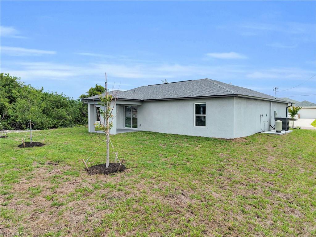 Rear exterior showcasing a covered patio with sliding glass doors, a gray shingle roof, light-colored stucco siding, and a spacious green lawn with newly planted trees