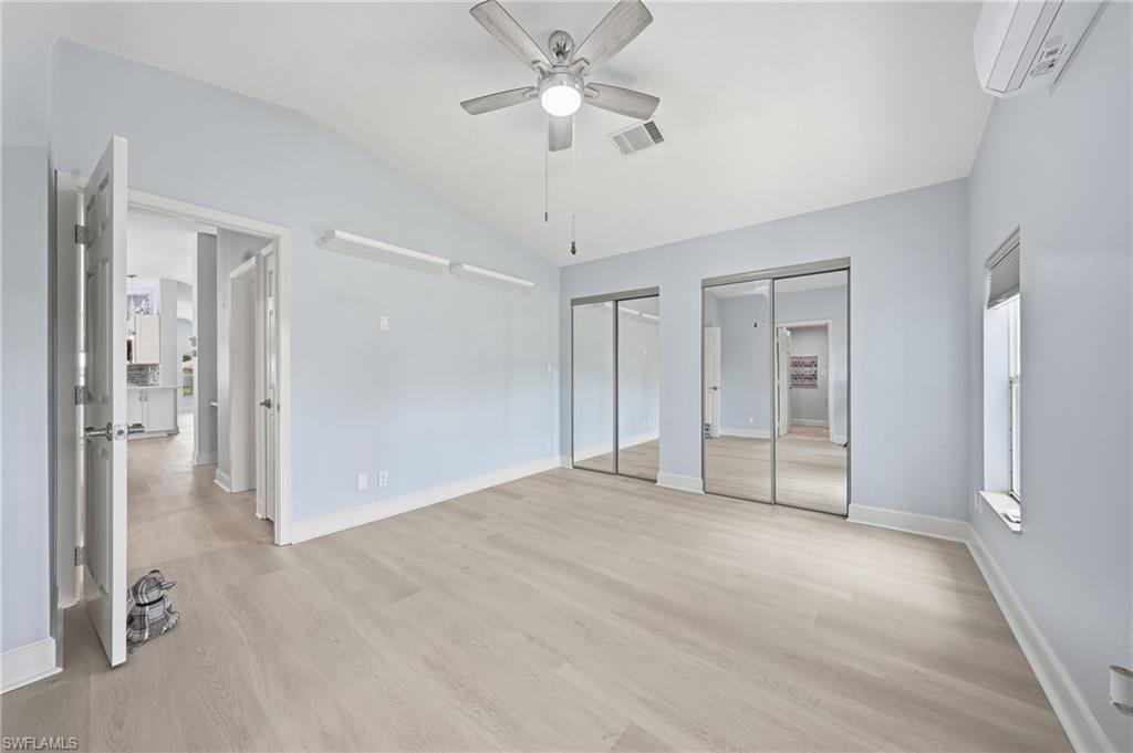 Spacious room featuring vaulted ceilings, light wood-finish flooring, two sets of mirrored sliding closet doors, a ceiling fan, and a wall-mounted mini-split AC unit