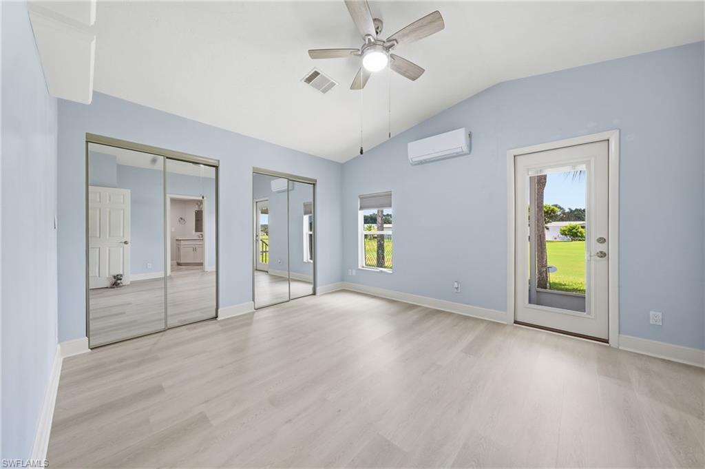 Spacious room featuring vaulted ceilings, light wood-finish flooring, two sets of mirrored sliding closet doors, a ceiling fan, and a glass-paneled exterior door