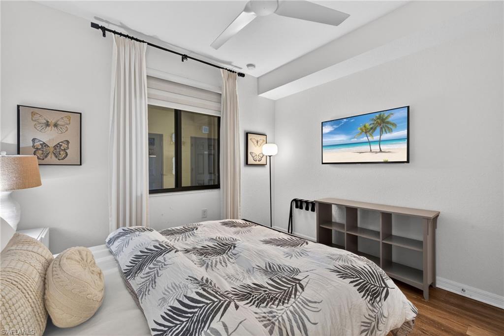 Bedroom featuring a large window with roller shades and full-height drapes, wood-finish flooring, a ceiling fan, and an integrated wall-mounted television