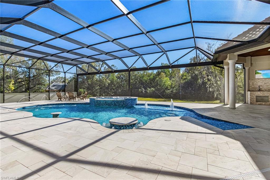 Expansive screened enclosure featuring a freeform swimming pool with integrated spa and water features, surrounded by light-toned tile decking, and an outdoor kitchen area with stone facing