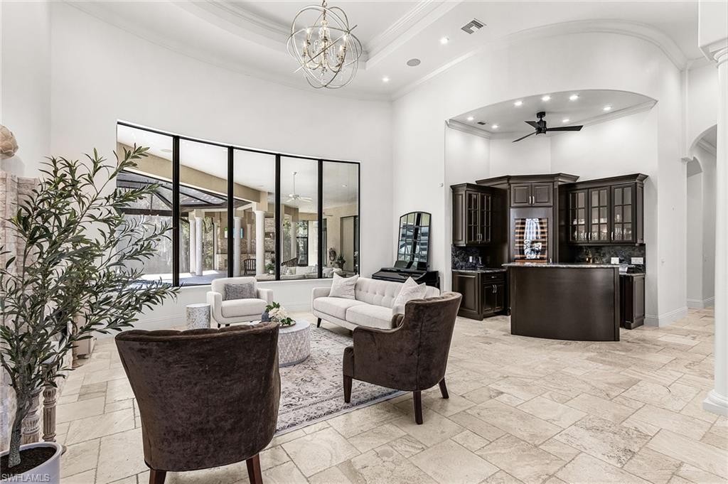 Curved floor-to-ceiling windows with black framing, high vaulted ceilings, large rectangular tile flooring, a built-in wet bar with dark wood cabinetry and stone countertops, and an ornate chandelier
