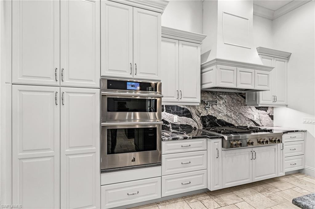 Kitchen featuring white cabinetry, stainless steel double wall ovens, a black and white veined backsplash, a professional-grade gas range, and stone tile flooring