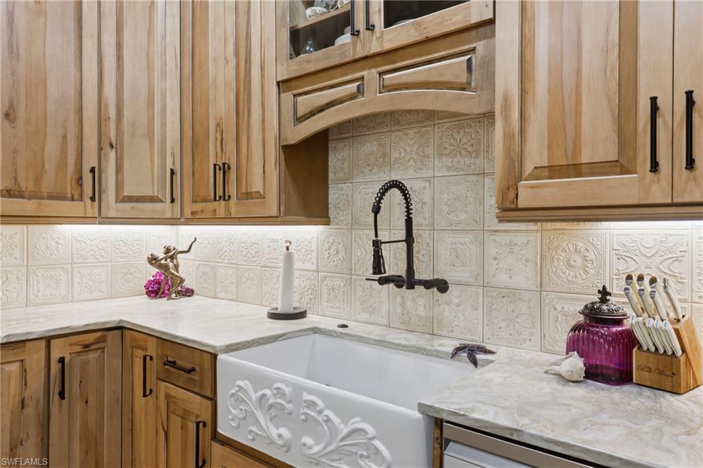 Farmhouse-style kitchen featuring an ornate apron sink with an oil-rubbed bronze wall-mounted faucet, light wood cabinetry, stone countertops, and a decorative tile backsplash