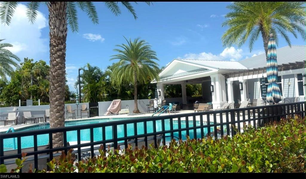 Resort-style swimming pool area featuring a black metal fence, mature palm trees, and a covered patio with white columns and a gray metal roof