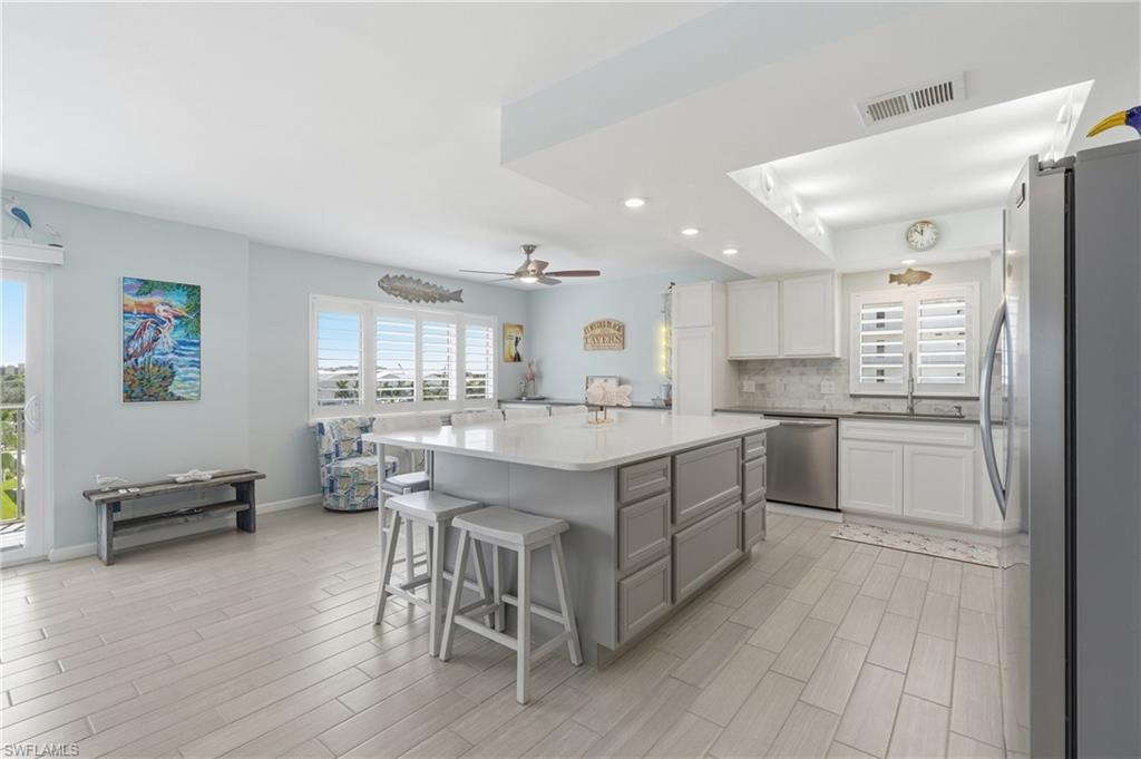 Open-concept living area with plank tile flooring, a kitchen island with seating, white cabinetry, stainless steel appliances, and plantation shutters - Virtually Edited Image