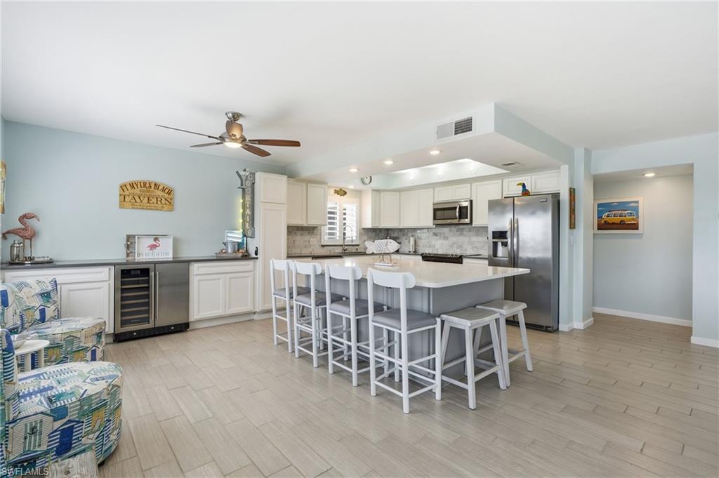 Open-concept kitchen and dining area featuring stainless steel appliances, white cabinetry, quartz countertops, a large island with seating for multiple guests, wood-finish flooring throughout, and ceiling fan with recessed lighting - Virtually Edited Image