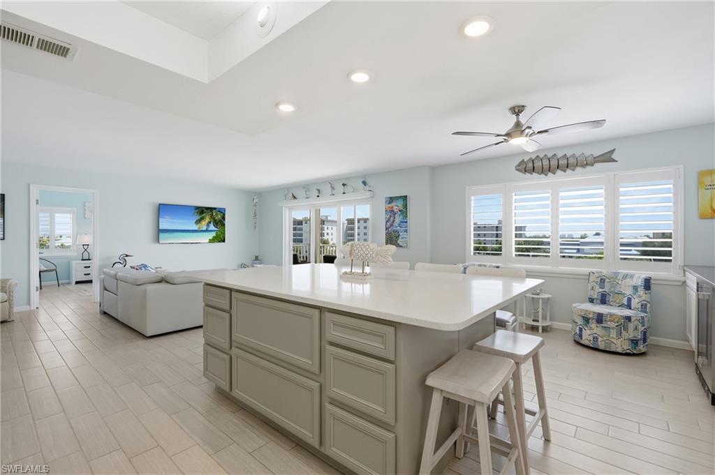 Open-concept living area featuring a large kitchen island with cabinetry in soft neutral tones and white quartz countertop, tile flooring throughout, recessed ceiling lighting, ceiling fan, and multiple glass doors and shuttered windows providing natural light and water views - Virtually Edited Image
