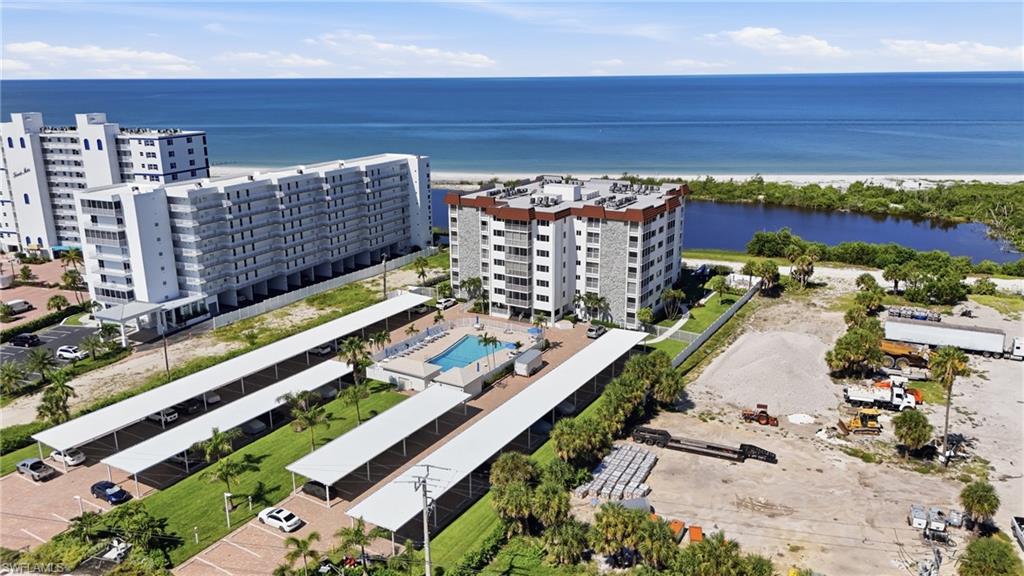 Modern waterfront community featuring contemporary multi-story residential buildings with ocean views, resort-style amenities including an expansive pool deck with lounging areas, manicured landscaping with mature trees, and direct access to coastal waterways and natural preserve areas - Virtually Edited Image