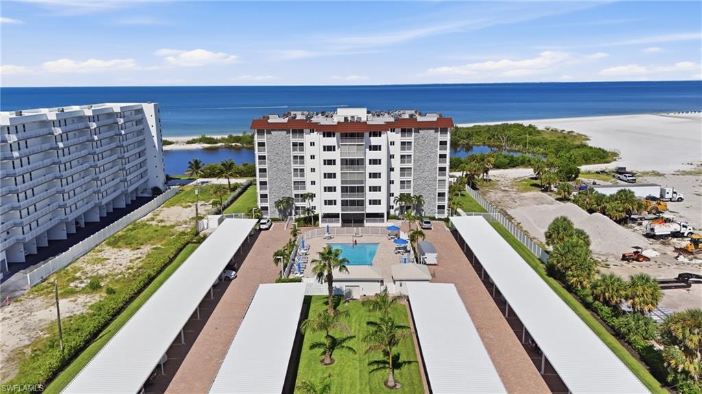 Waterfront residential community featuring a six-story apartment building with brick and stone exterior, resort-style amenities including a heated swimming pool, manicured landscaping with palm trees and green space, and direct access to sandy beachfront with unobstructed ocean views - Virtually Edited Image