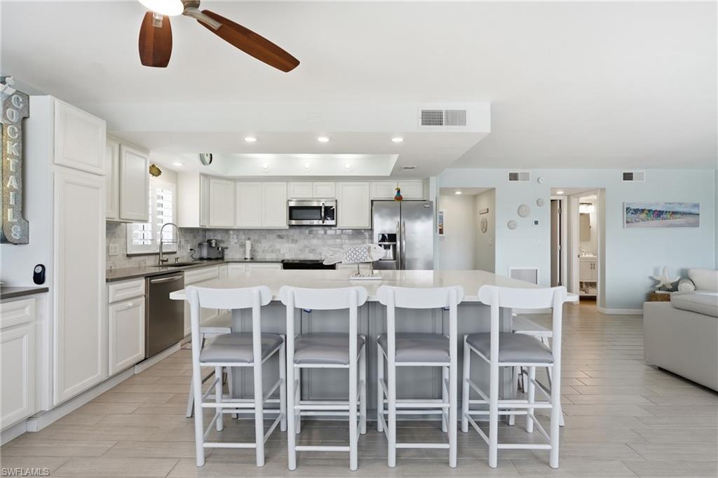 Open-concept kitchen and dining area featuring white cabinetry, stainless steel appliances, quartz countertops, a large center island with seating for five, wood-finish flooring throughout, and recessed ceiling lighting with a ceiling fan - Virtually Edited Image