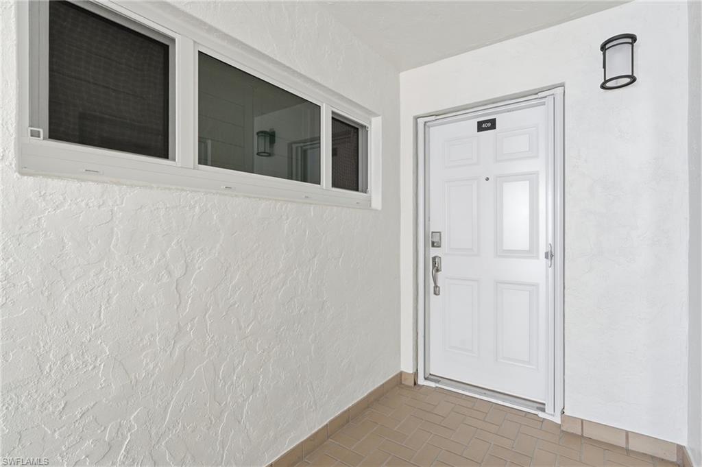 Bright entryway with white six-panel entry door, transom windows with dark screens, textured white walls, and warm wood-finish flooring - Virtually Edited Image
