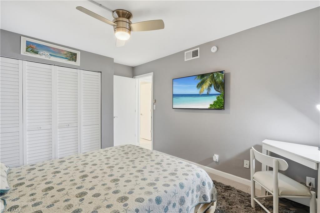 Bright secondary bedroom featuring neutral gray walls, patterned bedding, ceiling fan with integrated lighting, louvered closet doors, and a desk area with built-in shelving - Virtually Edited Image
