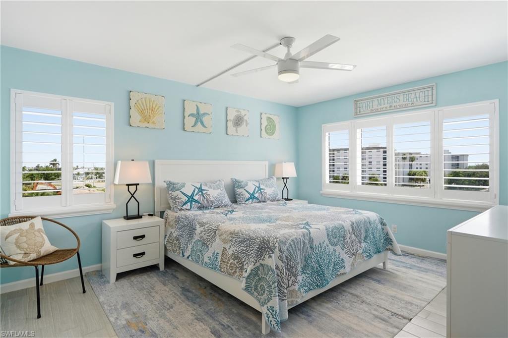 Serene coastal-inspired bedroom featuring soft turquoise walls, wood-finish flooring, and multiple windows with plantation shutters providing natural light and ventilation - Virtually Edited Image