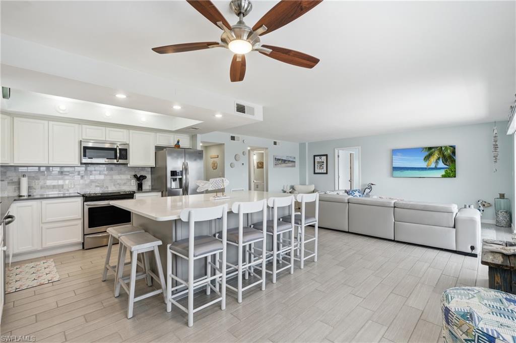 Open-concept kitchen and living area featuring white cabinetry, stainless steel appliances, quartz countertops, a large center island with seating for multiple, wood-finish flooring throughout, recessed and ceiling fan lighting, and sliding glass doors opening to additional living space - Virtually Edited Image