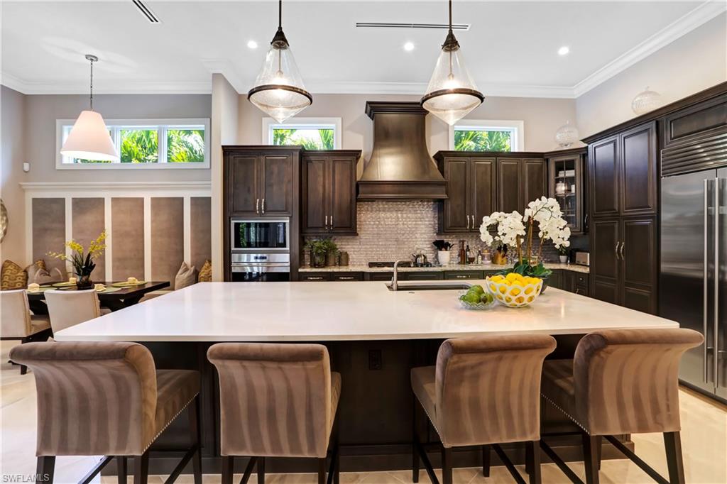 Spacious kitchen featuring a large central island with a white countertop, dark wood cabinetry, stainless steel appliances, a decorative range hood, and a tiled backsplash