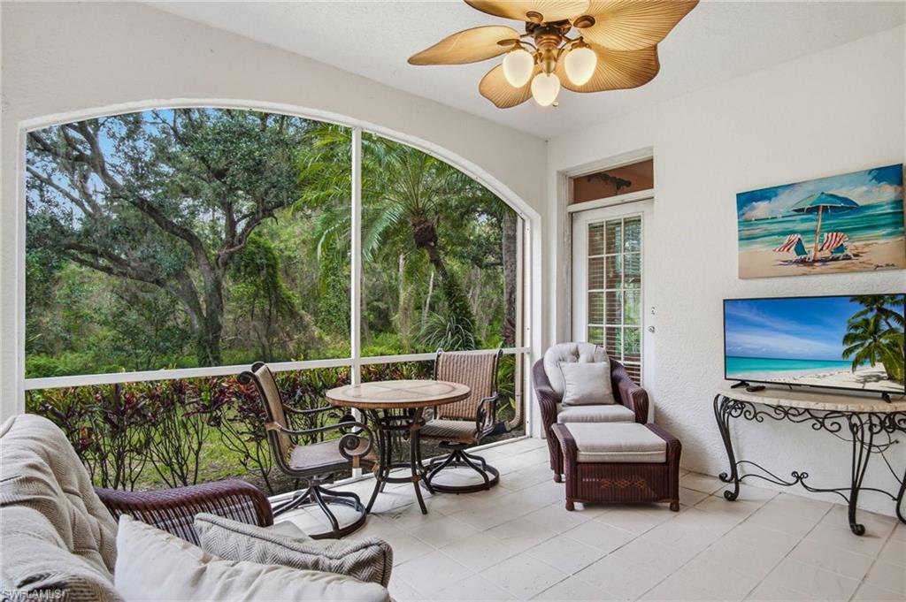 Expansive screened lanai showcasing panoramic windows framing mature oak trees, ceramic tile flooring, brass ceiling fan with integrated lighting, and multiple seating zones with built-in furnishings