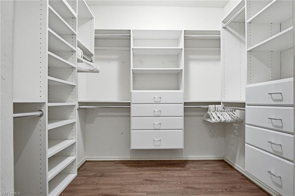 Walk-in closet featuring white built-in shelving, multiple hanging rods, four integrated drawers with silver pulls, and wood-finish flooring