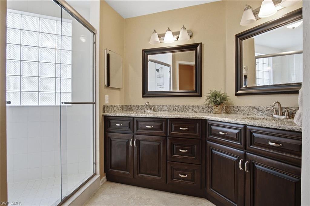 Shower enclosure with glass block window, dark wood-finish vanity with granite-style countertop, dual sinks, and two framed mirrors with vanity lighting