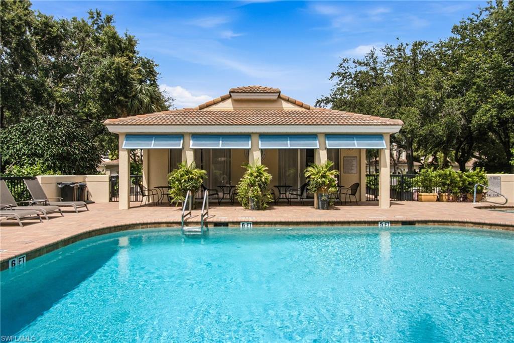 Outdoor swimming pool surrounded by a brick paver deck, featuring a pool house with a tiled roof and covered patio, and lush mature trees providing natural privacy