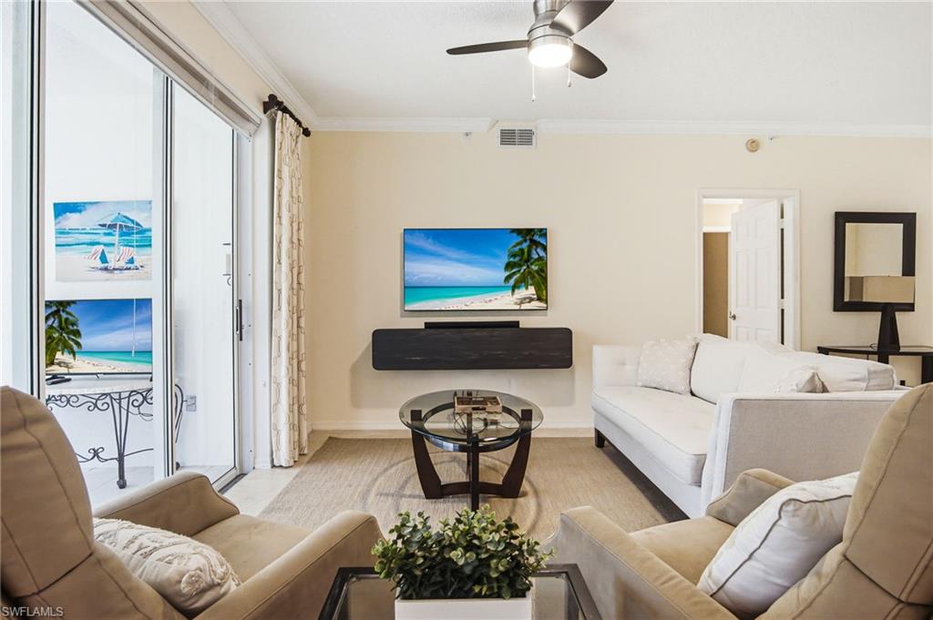 Bright, light-filled living space featuring floor-to-ceiling sliding glass doors with ocean vistas, neutral cream-toned walls, ceiling fan with integrated lighting, wall-mounted flat-screen television above a dark media console, comfortable seating arrangements, and a separate sleeping area with white bedding and dark-framed mirror accent