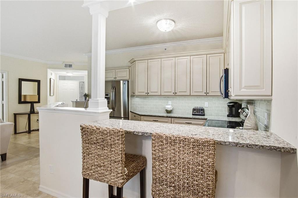 Bright, open-concept kitchen featuring white cabinetry with panel detailing, stainless steel appliances, and a large waterfall-edge granite island with seating for two