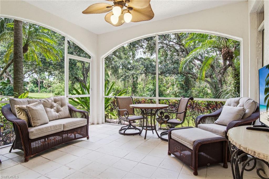 Expansive screened lanai featuring curved architectural detailing, floor-to-ceiling impact-resistant windows framing lush tropical landscaping, cream tile flooring, and integrated seating areas with woven rattan furnishings and bronze ceiling fan