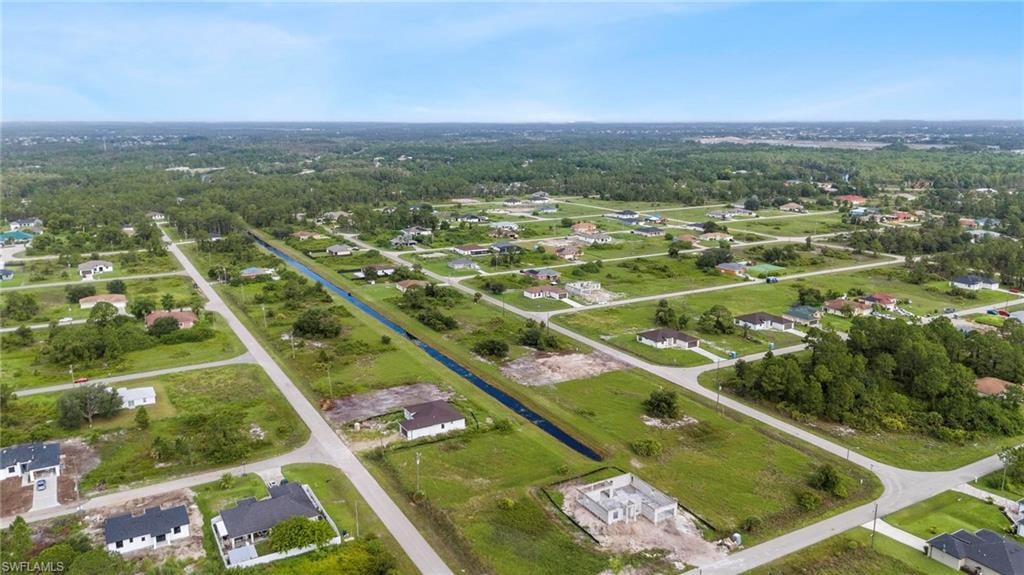 Aerial perspective of a developing residential area featuring multiple single-family homes, undeveloped lots, a central canal, paved streets, and abundant green space