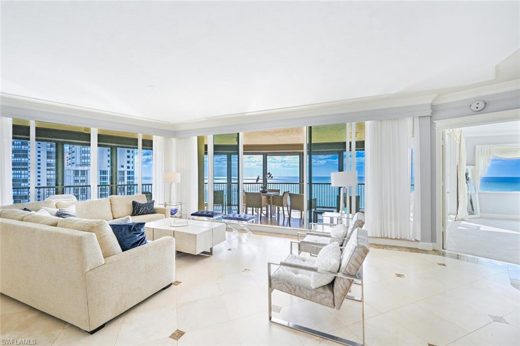 Spacious living area featuring large format tile flooring with accent inlays, full-height windows, a sliding glass door to an outdoor balcony, and a prominent ocean backdrop