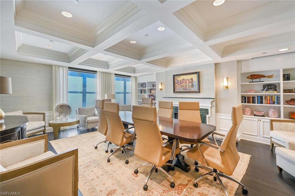 Spacious room featuring coffered ceilings, dark wood-finish flooring, built-in shelving, a fireplace with a stone surround, and large windows with privacy treatments