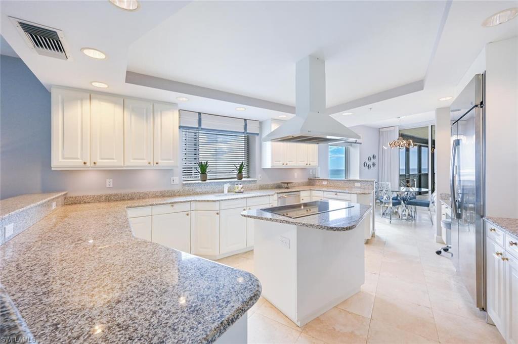 Modern kitchen featuring white cabinetry, light-colored stone countertops, a central island with a cooktop, a stainless steel refrigerator, and recessed lighting
