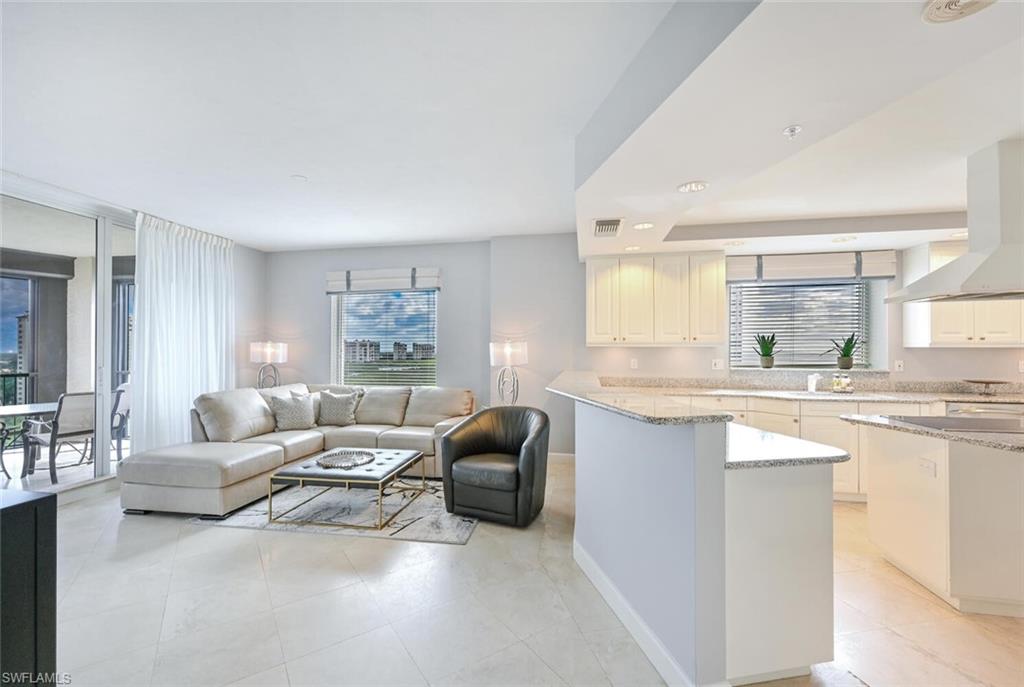 Open-concept living space featuring light-toned tile flooring, a kitchen with white cabinetry and light-colored countertops, and a living area with a large window and sliding glass doors opening to a balcony