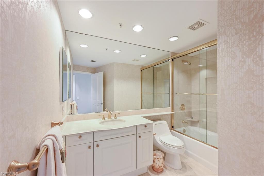 Bathroom featuring a white vanity with a light-colored countertop and a gold-toned faucet, a large wall-mounted mirror, recessed lighting, and a bathtub with a gold-framed glass enclosure