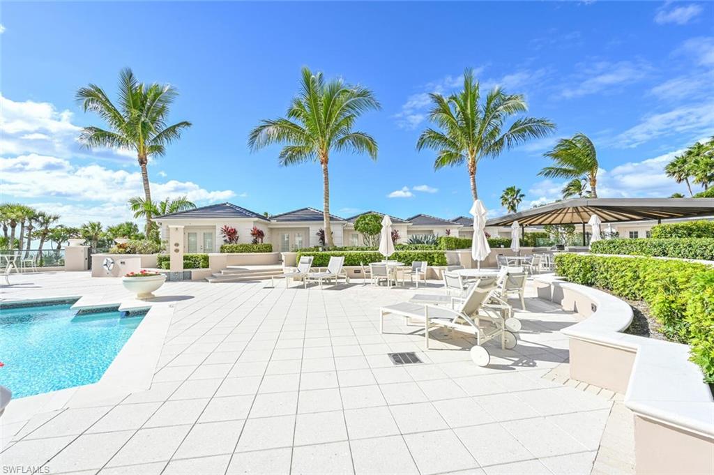 Expansive outdoor area featuring a swimming pool with a white tile surround, a large patio with light-colored pavers, lush green landscaping, and mature palm trees