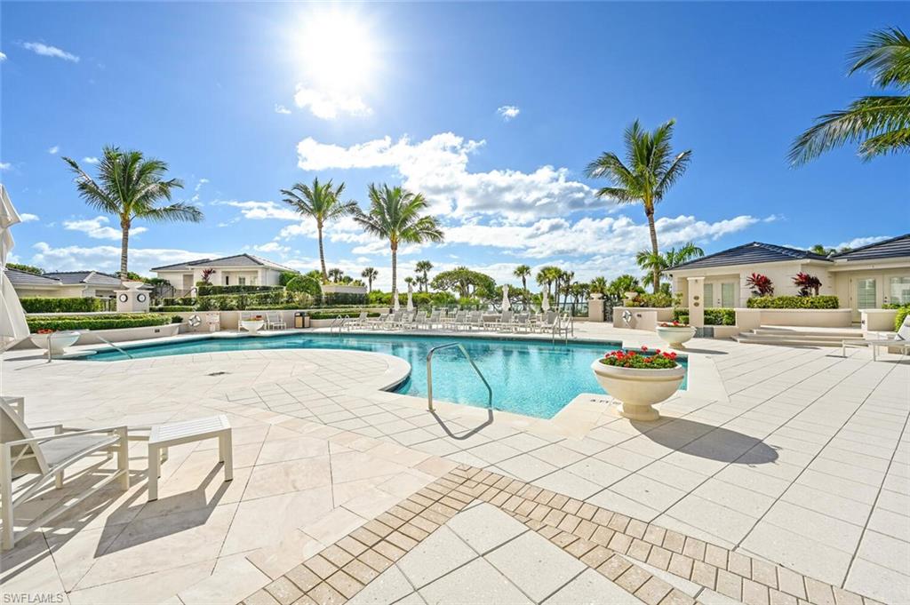 Spacious outdoor pool area featuring a freeform swimming pool, extensive paver decking, mature palm trees, lush landscaping, and surrounding structures with dark tile roofs