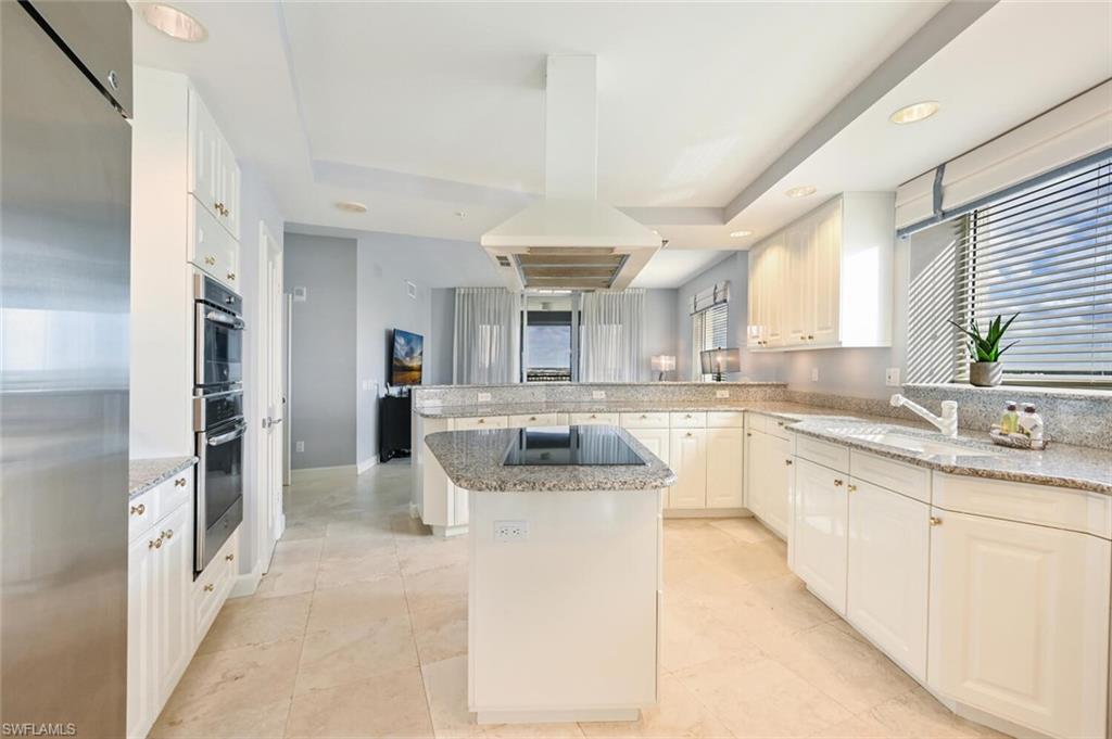Spacious kitchen featuring an island with a built-in cooktop, white paneled cabinetry, light-toned stone-finish countertops, stainless steel double wall ovens, and recessed lighting