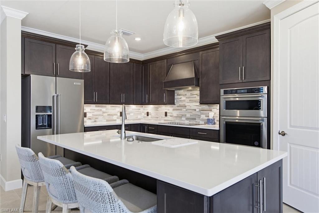 Kitchen featuring dark wood finish cabinets, stainless steel appliances, a center island with sink, decorative light fixtures, and light stone counters