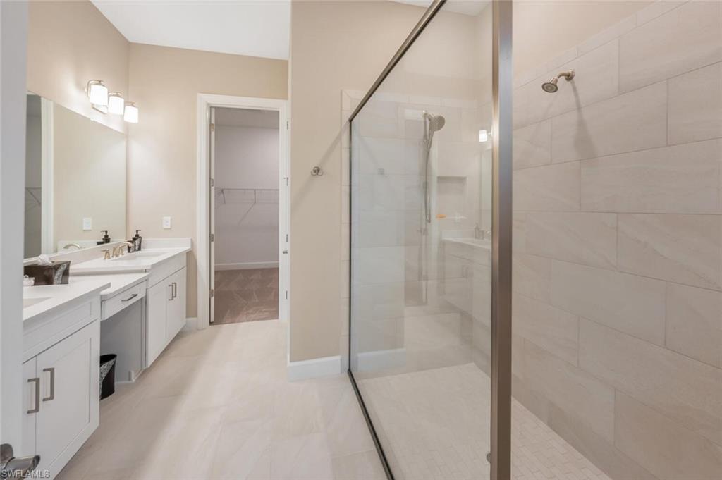 Spacious bathroom featuring a frameless glass shower enclosure, large format wall tiling, dual vanity with white cabinetry, light-toned countertop, and tile flooring