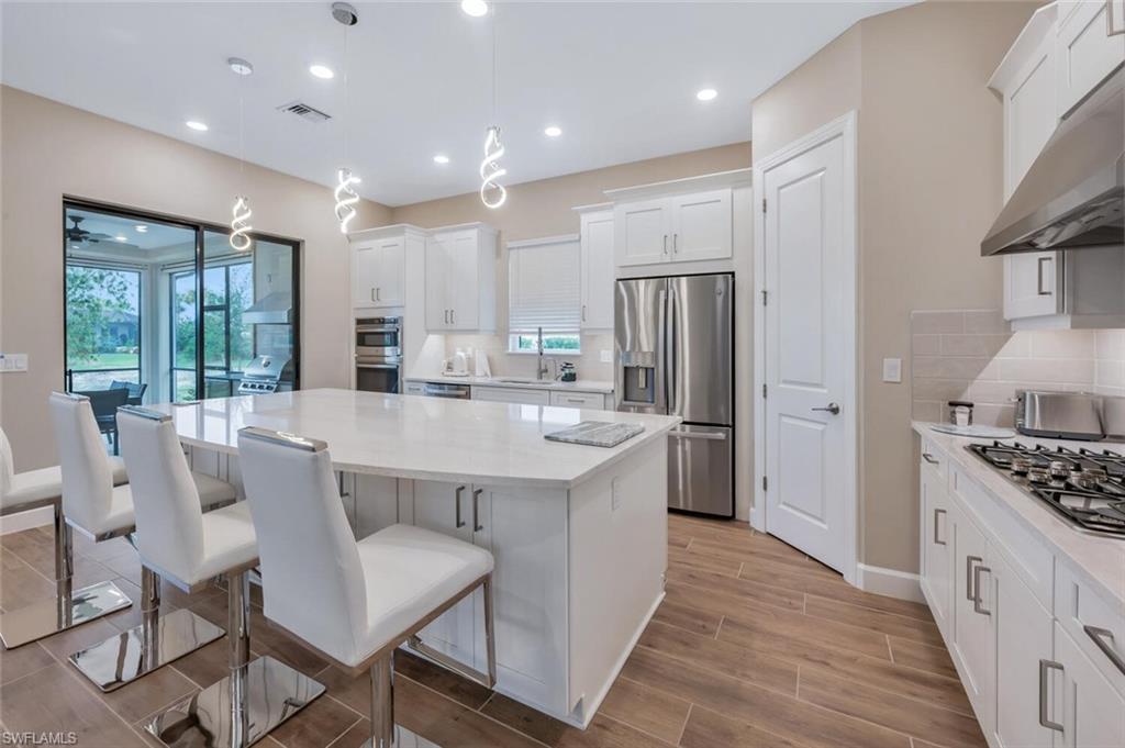 Open-concept kitchen featuring an expansive central island with ample seating, white shaker-style cabinetry, wood-finish tile flooring, recessed lighting, and stainless steel appliances