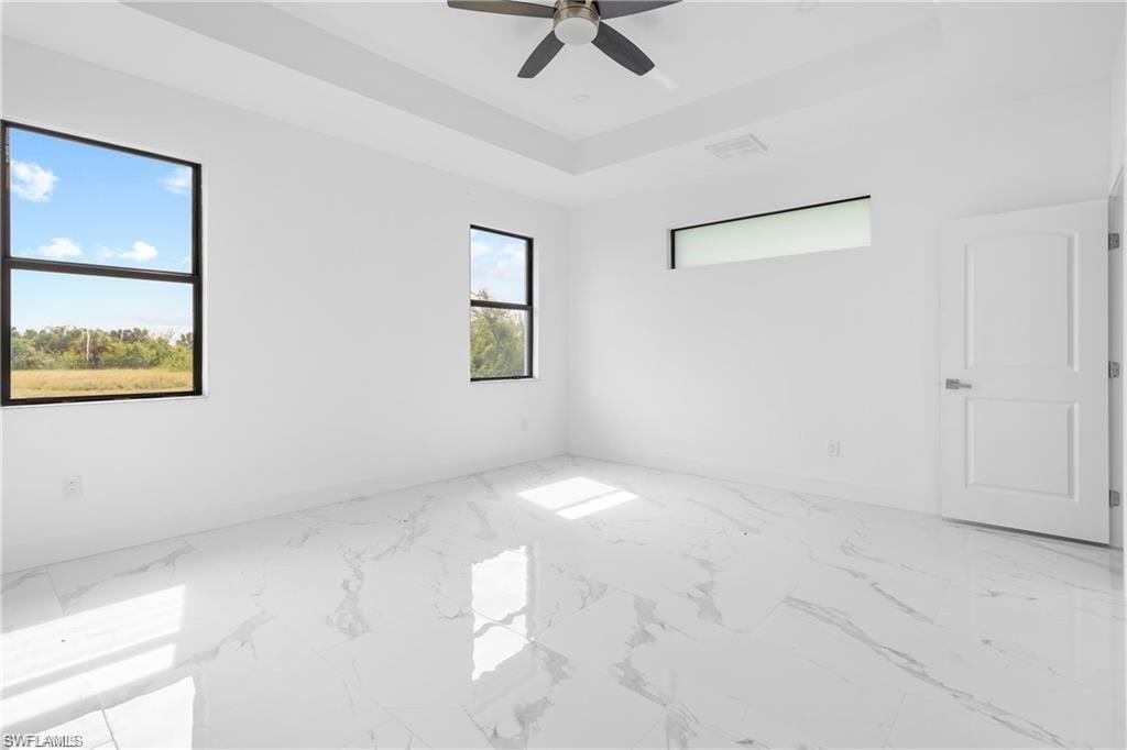 Spacious room featuring large format tile flooring, multiple windows providing natural light, a contemporary ceiling fan, and a tray ceiling