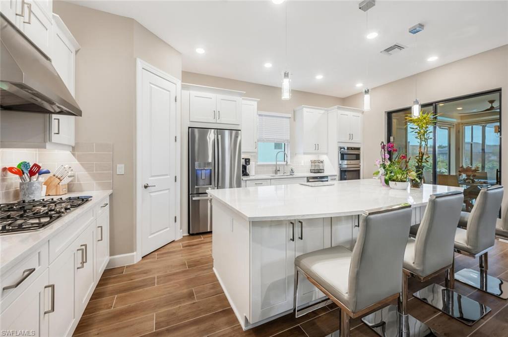 Open-concept kitchen featuring an expansive central island with seating, white shaker cabinetry, stainless steel appliances, and wood-finish flooring