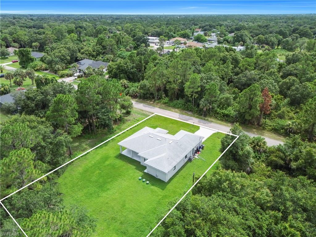Aerial property view featuring a light-colored roof, a perimeter outlined by white lines, a substantial green lawn, and surrounding mature trees